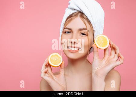 Portrait of an attractive cheerful woman with a towel wrapped around her head, holding lemon slices over pink background. Natural beauty Stock Photo