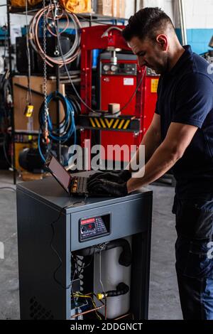 Adult man in using laptop to program broken machine during work in modern workshop Stock Photo