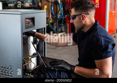 Adult man in goggles using laptop to program broken machine during work in modern workshop Stock Photo