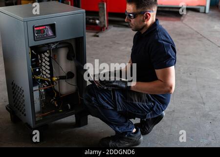 Adult man in goggles using laptop to program broken machine during work in modern workshop Stock Photo