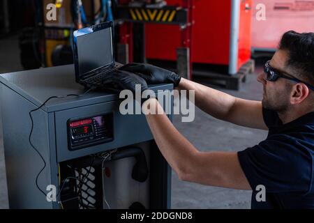Adult man in goggles using laptop to program broken machine during work in modern workshop Stock Photo