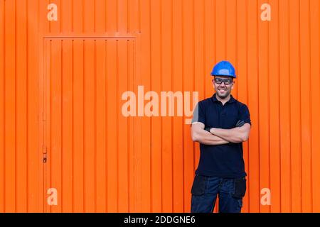 Positive confident adult male workman in blue protective hardhat and eyeglasses smiling friendly while standing against bright orange wall of workshop Stock Photo
