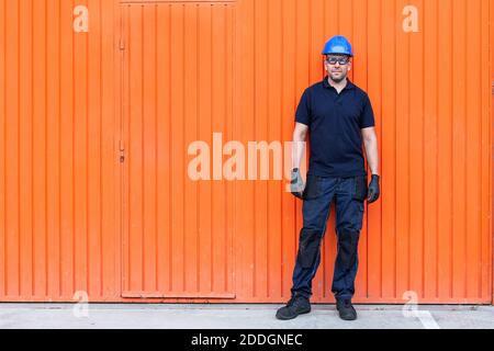 Positive confident adult male workman in blue protective hardhat and eyeglasses while standing against bright orange wall of workshop Stock Photo