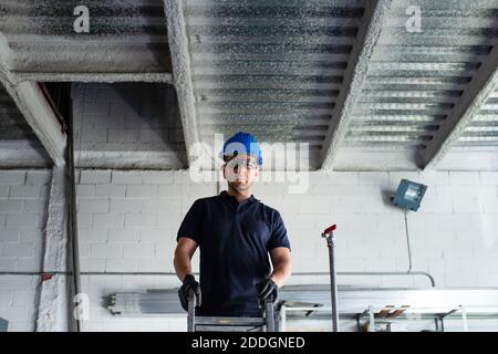 Low angle of cheerful male worker in hardhat and protective glasses standing on ladder and installing metal pipe while working in workshop Stock Photo