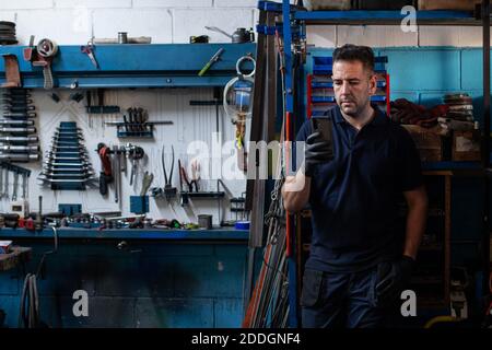Serious adult male mechanic in workwear standing near shelves with professional tools and instruments and checking information on smartphone while wor Stock Photo