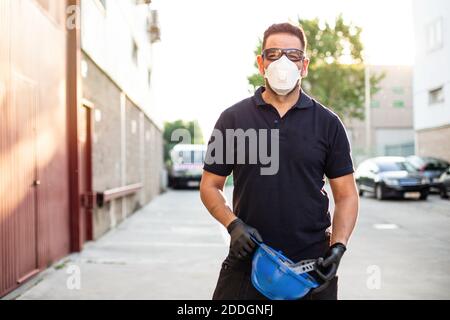 Adult male mechanic in protective glasses and respirator holding blue hardhat in hands and looking at camera while standing near workshop building on Stock Photo