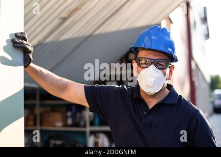 Adult male mechanic in protective glasses and respirator with blue hardhat looking at camera while standing near workshop building on street Stock Photo