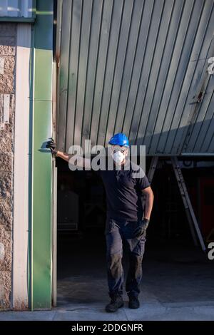 Adult male mechanic in protective glasses and respirator with blue hardhat looking at camera while standing near workshop building on street Stock Photo