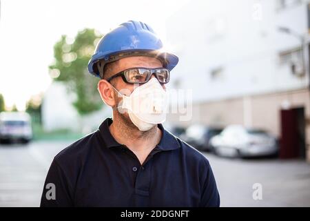 Adult male mechanic in protective glasses and respirator with blue hardhat looking away while standing near workshop building on street Stock Photo