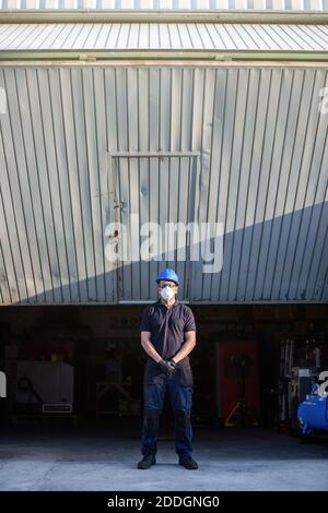 Adult male mechanic in protective glasses and respirator with blue hardhat looking at camera while standing near workshop building on street Stock Photo