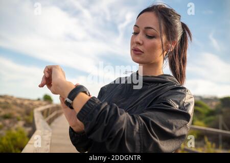 Active young brunette in black sports outfit standing on wooden pathway and checking data on fitness tracker after training in nature Stock Photo