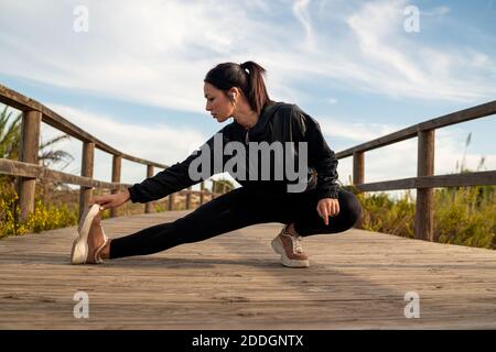 Full body of determined sportive fit female in black activewear doing stretching exercise while warming up before running on wooden walkway in nature Stock Photo