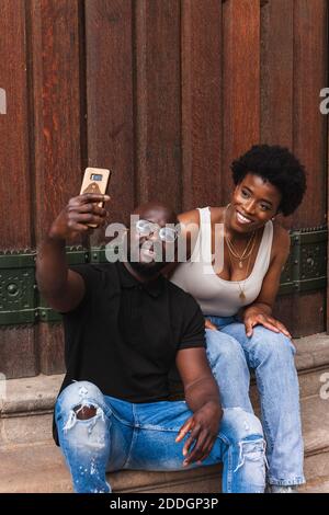 Cheerful African American couple in casual clothes sitting on stairs and taking selfie on smartphone Stock Photo