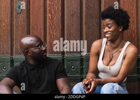 Amazed young African American female reacting on news from boyfriend while sitting together against wooden wall Stock Photo