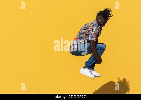 Full body side view of positive active young African American guy in trendy colorful shirt and jeans jumping high against bright yellow wall Stock Photo