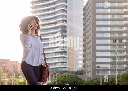 Cheerful ethnic female entrepreneur standing in downtown and having conversation on mobile phone while looking away Stock Photo