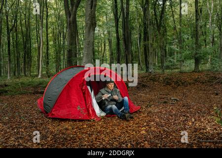 Calm male explorer sitting near tent and pouring hot drink from thermos in cup while enjoying camping in forest Stock Photo