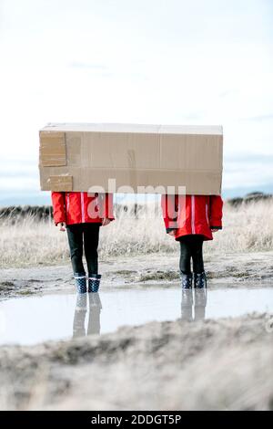 Anonymous children in rubber boots and raincoats standing in puddle and hiding behind cardboard package while having fun during weekend Stock Photo