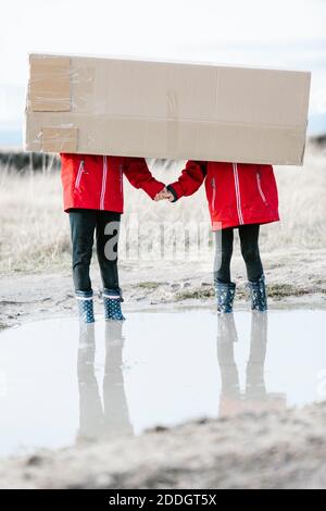 Anonymous children in rubber boots and raincoats standing in puddle and hiding behind cardboard package while having fun during weekend Stock Photo