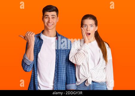 Surprised man smiles and points finger to free space, amazed female with open mouth presses hand to her cheek Stock Photo