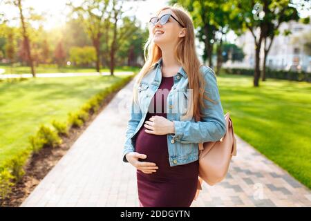 A beautiful pregnant girl student, in sunglasses, happily looks at the sky, walks in the park after studying. Stock Photo