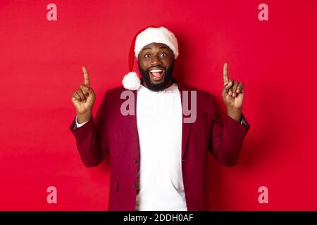 Christmas, party and holidays concept. Excited Black man in santa hat and blazer, pointing fingers up and smiling, showing promo offer, red background Stock Photo
