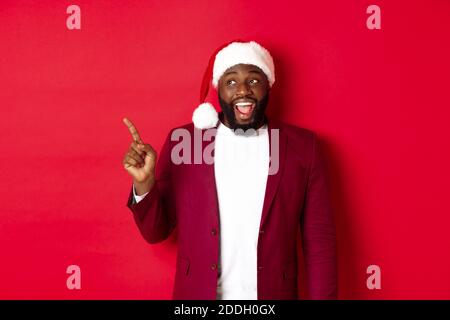 Christmas, party and holidays concept. Cheerful Black guy in santa hat pointing fingers left, showing banner, standing amused against red background Stock Photo