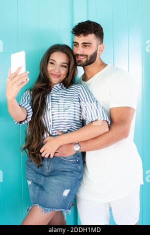 Young couple leaning on a green door and taking a selfie. They are both smiling and he is hugging her. Stock Photo