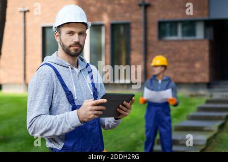 Portrait of young male builder looking at camera while holding digital tablet pc with coworker working in the background at cottage construction site. Selective focus Stock Photo