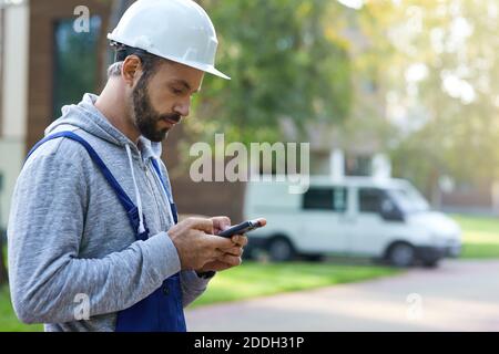 Young male builder in hardhat using smartphone, standing outdoors while working at cottage construction site. Profession, construction and building concept. Side view Stock Photo
