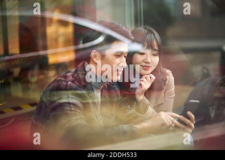 through the glass shot of a young asian couple sitting on ground looking at cellphone Stock Photo