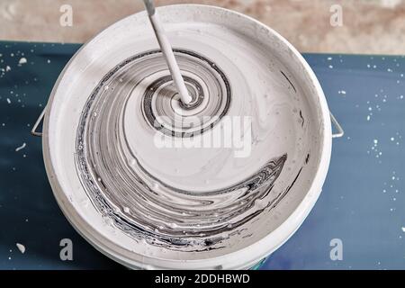 Handyman is mixing black and white colors using painting mixer in the basket to get a gray color. Worker mixes paints with help of drill. Stock Photo