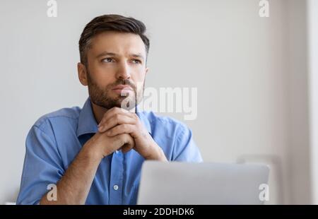 Portrait Of Pensive Businessman Sitting At Workplace With Laptop And Looking Away Stock Photo