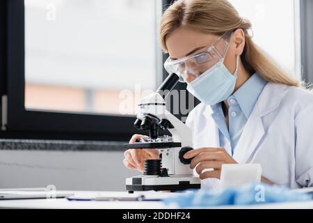 scientist in medical mask and goggles looking through microscope Stock Photo