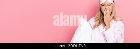 Young woman in pajamas and blindfold holding pillow and yawning on pink background, banner Stock Photo