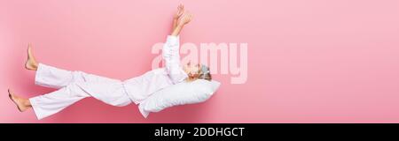 Young woman in pajamas and blindfold lying on pillow on pink background, banner Stock Photo