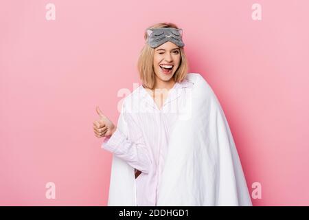 Cheerful woman in pajamas and blanket winking and showing like on pink background Stock Photo