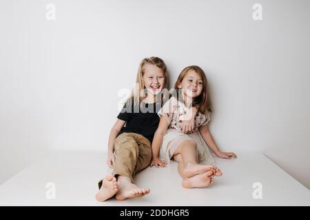 Smiling siblings sitting together on table. Boy hugs girl, she leans away a bit. Stock Photo