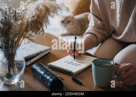 2021 goals, new year resolution, planning. Woman writing in Notebook with text 2021 loading on the table in apartments with cat. Notepad list concept Stock Photo