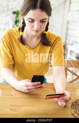 Cute young woman in bright yellow T-shirt sits at table and makes purchases online using smartphone and credit card. Shopping online during the corona Stock Photo