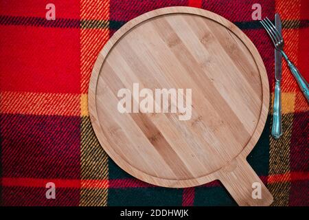 Cold season menu background, cooking or baking: round empty wooden cutting board on checkered cloth with knife and fork Stock Photo
