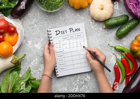 Woman Filling Weekly Diet Plan At Table With Composition Of Fresh Vegetables Stock Photo