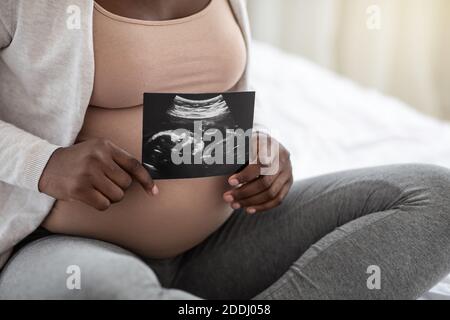 Pregnant black woman sitting on bed holding ultrasound image of baby, closeup Stock Photo