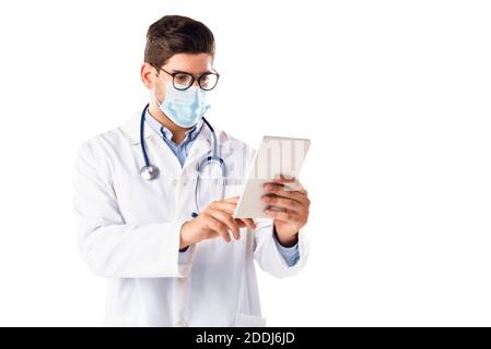 Portrait shot of male doctor wearing face mask and using digital tablet while standing at isolated white background. Stock Photo