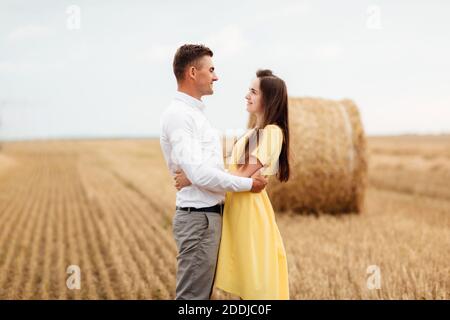 Photo of joyful couple man and woman walking through golden field with bunch of haystacks and hugging together during sunny day. Stock Photo