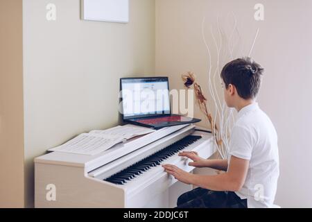 12 year old boy playing the white piano looking at the notes of his laptop. Stock Photo