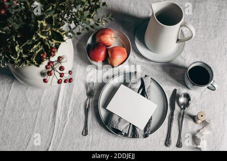 Festive Christmas table setting. Silver cutlery, bowl of apples on linen tablecloth. Cup of coffee, pitcher. Defocused bouquet of holly berries, rose Stock Photo
