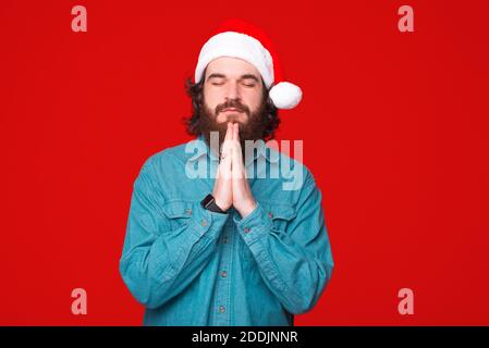 Young bearded man wearing Christmas hat is holding his hands praying over red background. Stock Photo