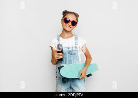 Happy teenager, girl in sunglasses with a skateboard and a refreshing drink in her hands. Posing on a white background. Stock Photo