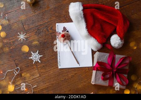 Flatlay on wooden brown floor New Year's mood. Writing Christmas wishes to Santa Claus. On textured background lies notepad in cage, deer pompom pen, Stock Photo